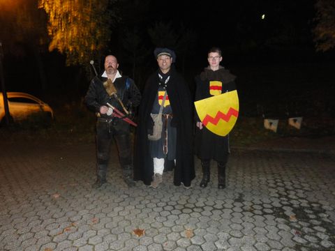 Drei Männer in historischen Kostümen stehen in einer nächtlichen Umgebung. Einer trägt ein Schild, während die anderen Waffen halten.