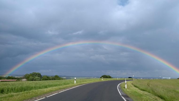 Eine kurvenreiche Straße unter einem bunten Regenbogen. Der Himmel ist bewölkt, mit grünen Wiesen auf beiden Seiten.