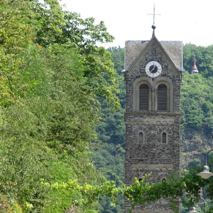 Ein Turm mit einer Uhr und einem Kreuz, umgeben von Bäumen und sanften Hügeln. Die Architektur ist historisch und harmoniert gut mit der Natur.