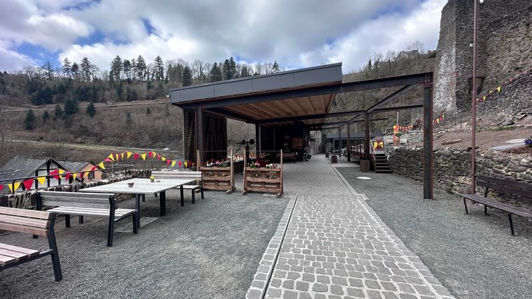 Ein modernes Restaurantgebäude mit einer gepflasterten Terrasse inmitten einer natürlichen Umgebung. Im Hintergrund sind Ruinen und farbige Fahnen zu sehen.