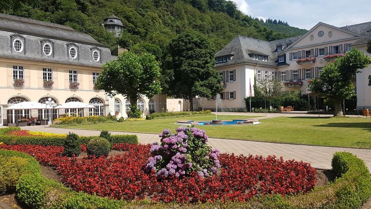 Ein schöner Garten mit bunten Blumenbeeten und gepflegten Rasenflächen. Im Hintergrund sind elegante Gebäude und grüne Bäume zu sehen.