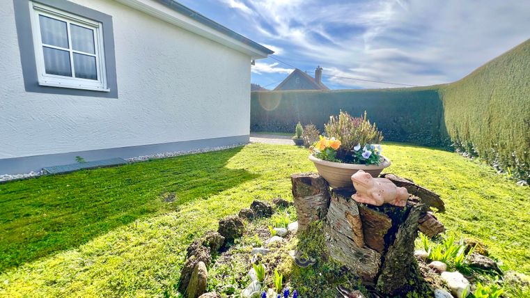 Ein gepflegter Garten mit einem Baumstumpf, der mit Blumen dekoriert ist. Im Hintergrund sieht man ein helles Haus und einen blauen Himmel.