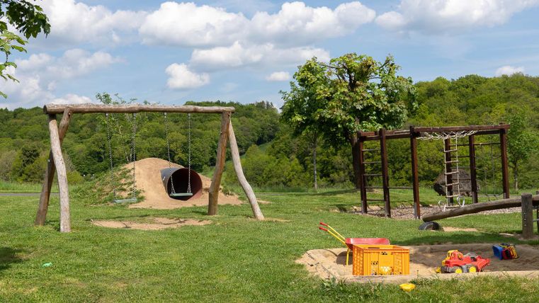 Ein Spielplatz mit einer Wippe, einer Rutsche und einer Sandfläche. Im Hintergrund sind grüne Bäume und ein blauer Himmel zu sehen.