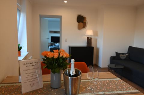 Ein gemütlicher Wohnbereich mit einem Tisch, auf dem eine Vase mit orangefarbenen Rosen steht. Im Hintergrund sind ein Sofa und ein Fernsehmöbel zu sehen.