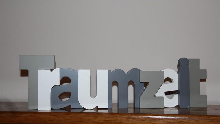 Eine dekorative Schriftzug aus bunten Buchstaben, die das Wort „Traumzeit“ bilden. Der Hintergrund ist schlicht und neutral.