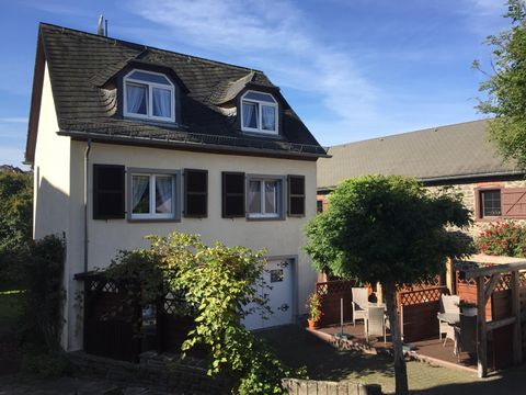 Ein zweigeschossiges Haus mit weißer Fassade und braunen Fensterläden. Im Vordergrund befindet sich ein Garten mit Möbeln und einem kleinen Baum.