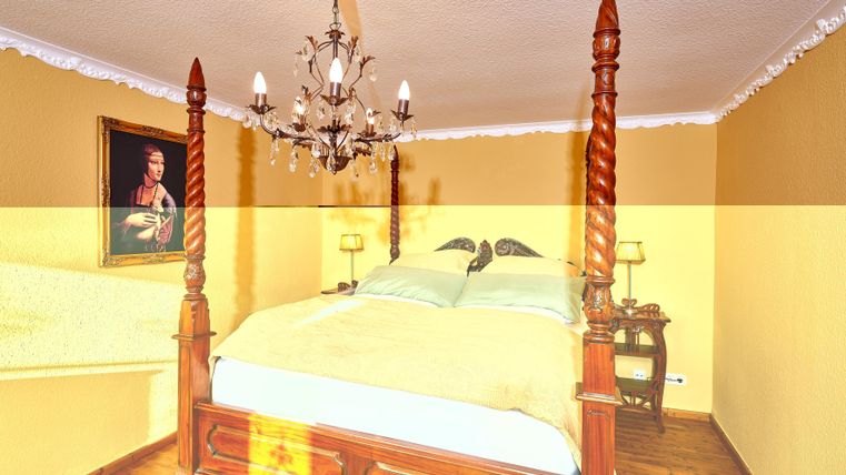 Ein gemütliches Schlafzimmer mit einem Himmelbett und einem eleganten Kronleuchter. Die Wände sind in warmem Gelb gestrichen und es gibt ein klassisches Gemälde an der Wand.