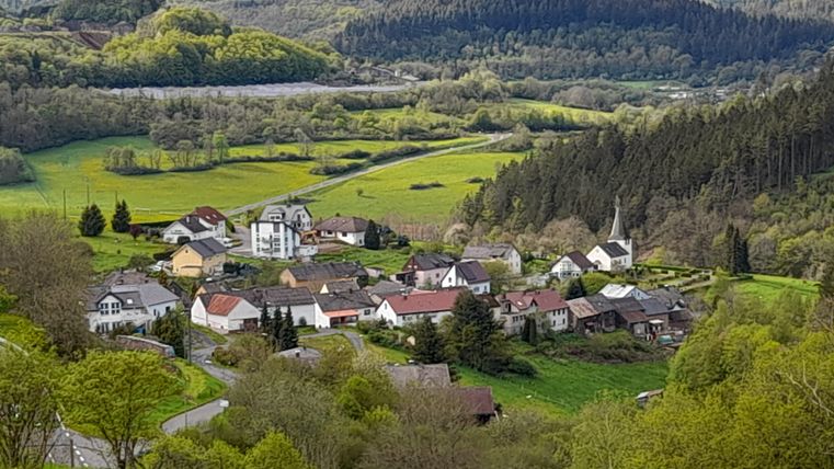 Ein malerisches Dorf umgeben von grünen Wiesen und Wäldern. Die Landschaft ist idyllisch und hügelig.