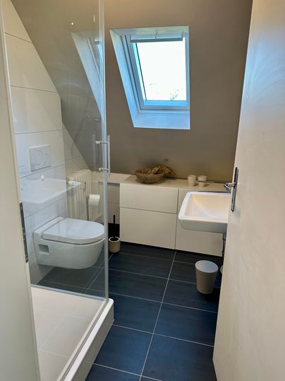 Ein modernes Badezimmer mit einer Dusche und einem Fenster. Die Wände sind in hellen Farben gehalten und der Boden ist mit dunklen Fliesen ausgestattet.