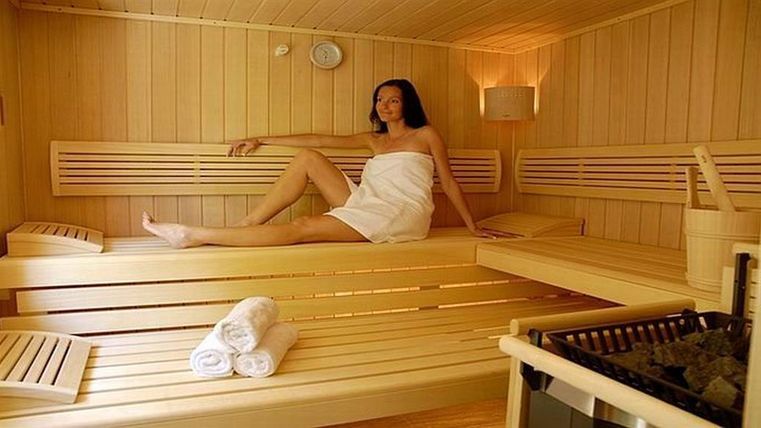 Eine gemütliche Sauna mit Holzverkleidung und sanftem Licht. Eine Frau sitzt entspannt auf einer Bank und genießt die Atmosphäre.