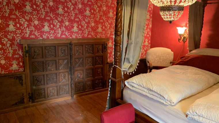 Ein gemütliches Schlafzimmer mit einer großen Himmelbett und einer roten Tapete. Im Hintergrund ist ein hölzerner Schrank und ein eleganter Sessel zu sehen.