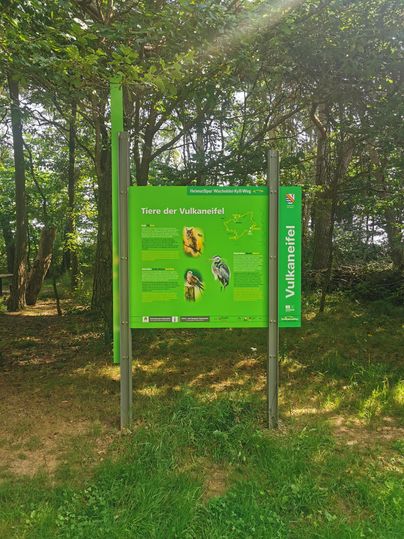 Ein Informationsschild über die Tiere der Vulkaninsel im Wald. Es zeigt Bilder und Namen von verschiedenen Tieren.