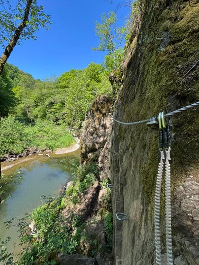 Ein schöner Wanderweg entlang eines Flusses, umgeben von üppigem Grün und hohen Felsen. Der Himmel ist klar und blau.