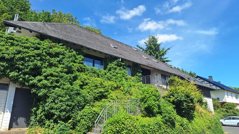 Ein gemütliches Haus, umgeben von grünen Pflanzen und Bäumen. Der Himmel ist blau mit einigen Wolken.
