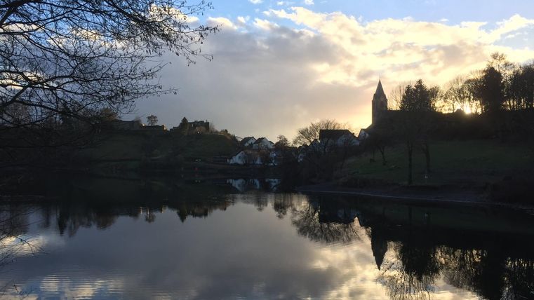 Ein ruhiger See mit spiegelndem Wasser und dramatischem Himmel. Am Ufer sind Bäume und ein Kirchturm sichtbar.