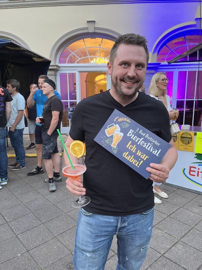 Ein lächelnder Mann hält ein Getränk in der Hand und zeigt ein Schild mit der Aufschrift "Bierfestival - ich war dabei!". Im Hintergrund sind andere Menschen auf einer Veranstaltung zu sehen.