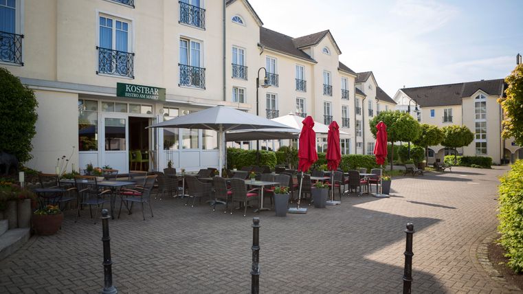 Eine einladende Terrasse mit roten Sonnenschirmen vor einem modernen Gebäude. Tische und Stühle laden zum Verweilen ein.