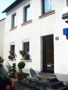 Ein weißes Wohnhaus mit braunen Fensterrahmen und einer dunklen Eingangstür. Vor dem Haus stehen Pflanzen in Töpfen.