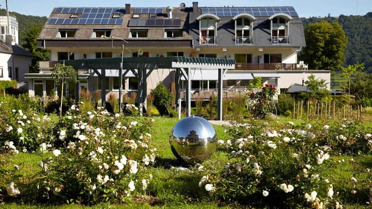 Ein modernes Gebäude mit Solarpanelen steht in einem schönen Garten. Der Garten ist mit zahlreichen Rosen und einer großen, glänzenden Kugel dekoriert.