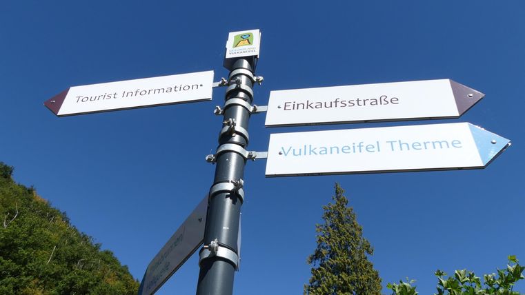 Ein Wegweiser mit mehreren Richtungen zeigt auf die Tourist-Information, die Einkaufsstraße und die Vulkan-Eifel-Therme. Der Himmel ist klar und blau.