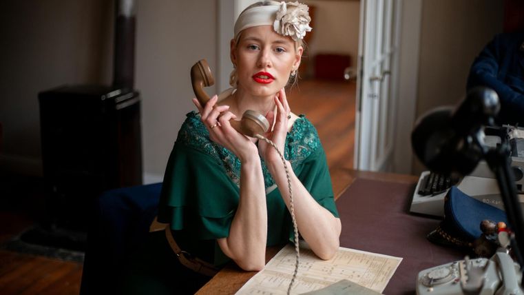 Eine elegante Frau mit rotem Lippenstift sitzt an einem Schreibtisch und hält einen Telefonhörer. Sie trägt ein grünes Kleid und hat eine Blumenschmuck im Haar.