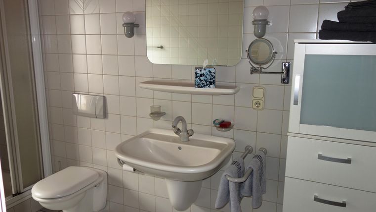 Ein modernes Badezimmer mit weißen Fliesen und einem Waschbecken. Im Hintergrund sind Dusche und Spiegel zu sehen.