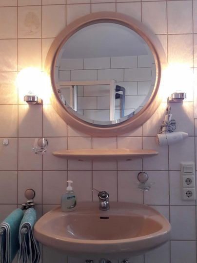 Ein Badezimmer mit einem runden Spiegel, einem Waschbecken und zwei Wandleuchten. Es gibt Handtücher und Pflegeprodukte auf einem Regal.