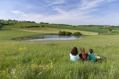 Drei Personen sitzen auf einer Wiese und schauen auf einen kleinen Teich. Die Landschaft ist grün mit sanften Hügeln und einem blauen Himmel.