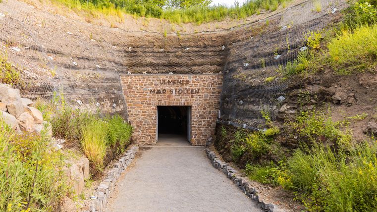Ein Eingang zu einer Höhle mit einer Steinfassade und einem beschrifteten Schild. Die Umgebung ist grün und hügelig, mit Gras und Bäumen.