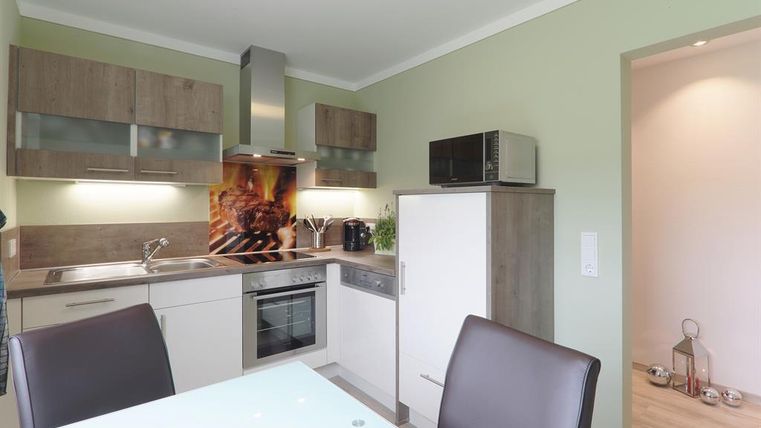 Eine moderne Küche mit Holzschränken und Edelstahl-Geräten. Der Raum ist hell und freundlich mit einem großen Tisch und Stühlen.