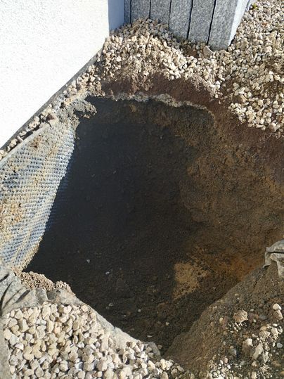 Ein großes Loch im Boden, umgeben von Erdmaterial und kleinen Steinen. An der Wand ist ein Netz sichtbar, das wahrscheinlich für Drainagezwecke dient.