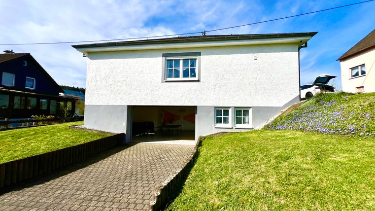 Ein modernes, zweigeschossiges Haus mit einer weißen Fassade und einem Garagenbereich. Der gepflegte Garten und der blaue Himmel schaffen eine freundliche Atmosphäre.