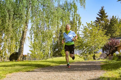 Ein älterer Mann joggt auf einem gepflasterten Weg in einer grünen Umgebung. Bäume und Sträucher umgeben die Szenerie unter einem klaren blauen Himmel.
